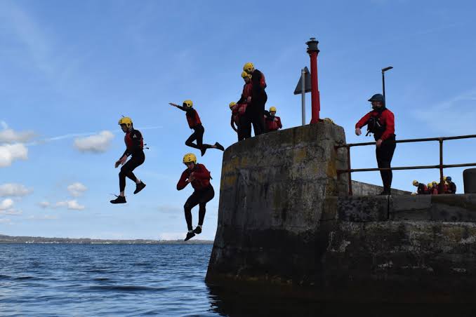 A group in wetsuits and helmets jumping off the pier into Carlingford Lough
                    on a coasteering session.