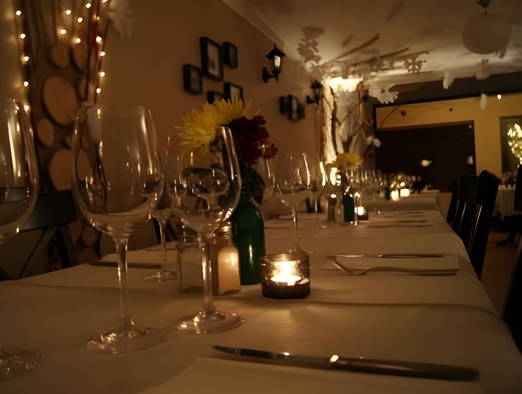 A candlelit table set for dinner at The Bay Tree: wine glasses, a small
                vase of flowers and a tealight down a long white cloth.