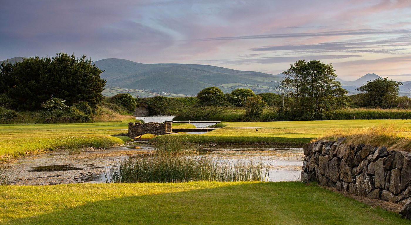 Greenore Golf Club at dusk: a stone bridge over the water hazard, fairway and
                    green beyond, with the mountains behind and a pink evening sky.