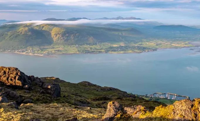 The view from the ridge above Carlingford: rocky outcrops in the foreground,
                    the marina below, and cloud spilling over the Mourne Mountains across the
                    lough.